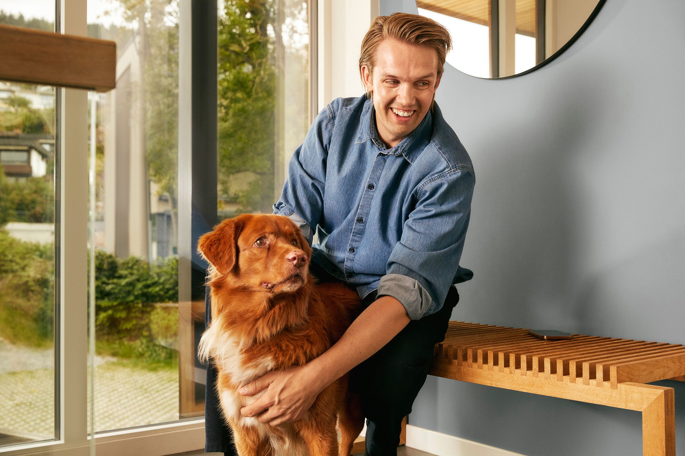 Smilende lyslugget mann med blå skjorte sitter på en benk og klapper en rødfarget hund