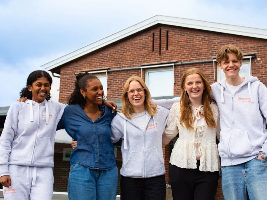 Fem smilende ungdommer foran skolebygning