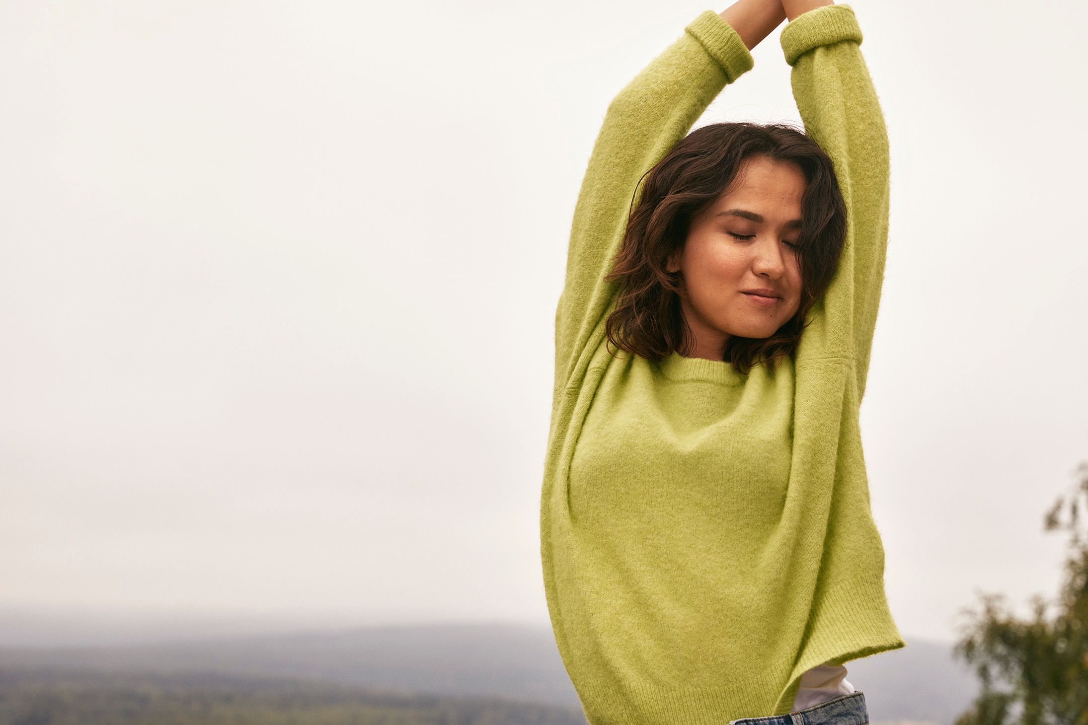 Ung kvinne i grønn genser og lukkede øyne strekker hendene samlet over hodet