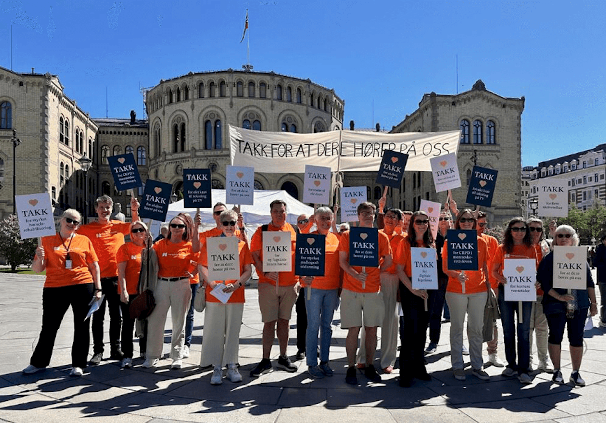 En stor gruppe mennesker med t-skjorter med Hørselsforbundet-logo står foran Stortinget med et banner og plakater med teksten Takk for at dere hører på oss.